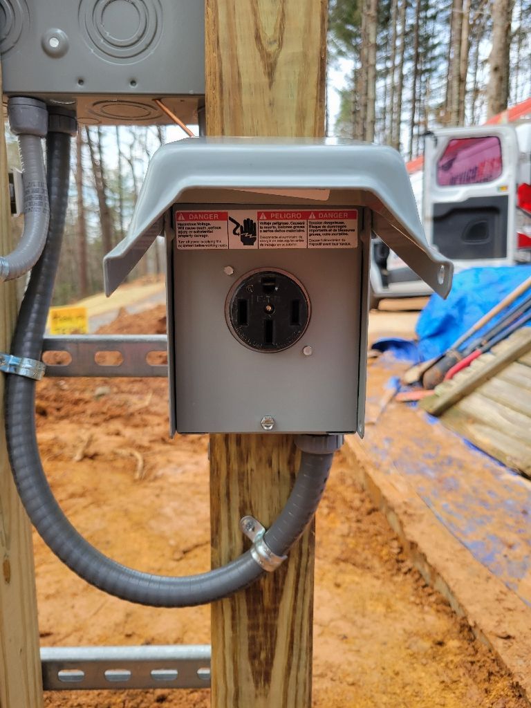 Outdoor electrical outlet mounted on a wooden post. Gray box with a black and gray outlet, a protective cover, and electrical conduit.