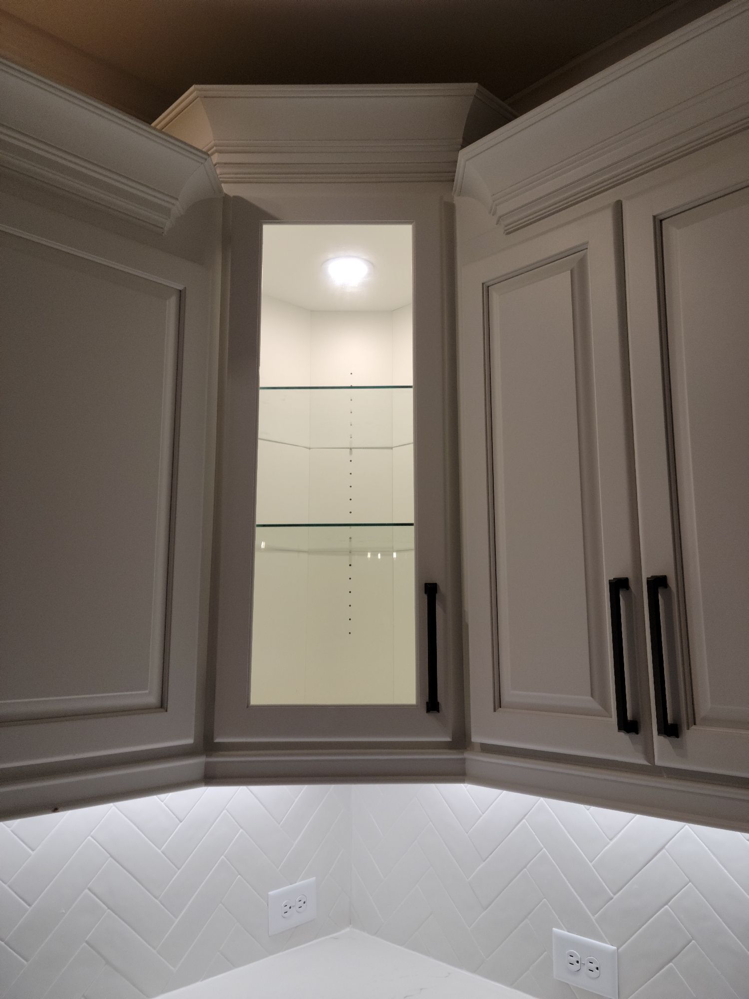 White kitchen cabinets with black handles, one glass-fronted and lit.