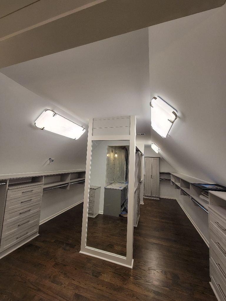 Walk-in closet with sloped ceiling, built-in shelves, large mirror, and wood flooring. Bright overhead lighting.