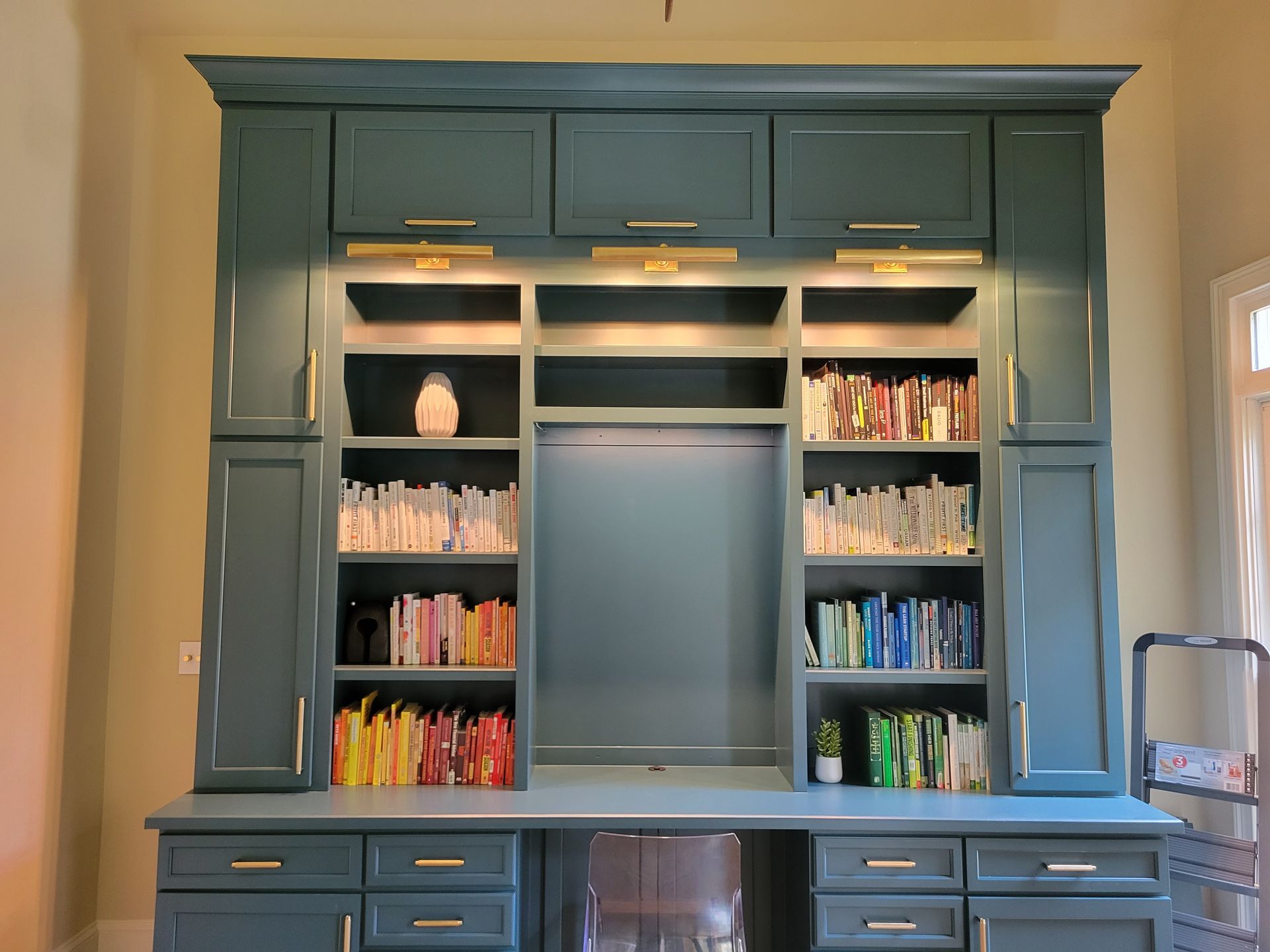 Blue built-in desk and bookshelves with overhead cabinets and gold hardware, brightly lit.