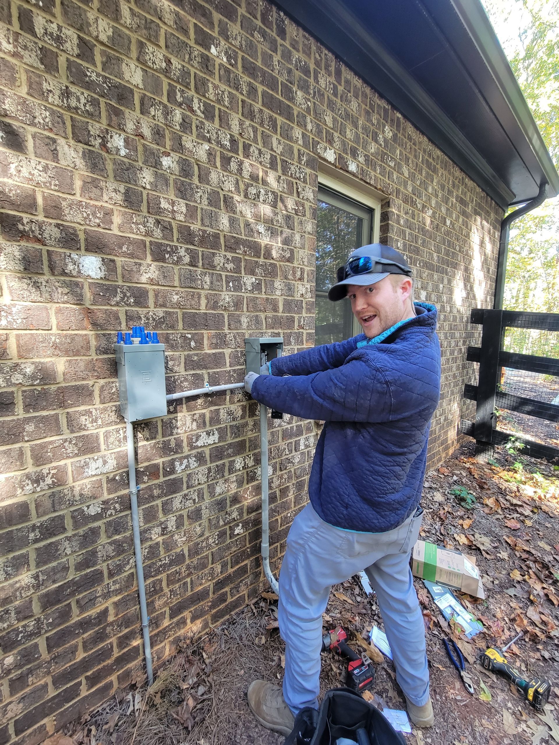 Man working on electrical box on brick building exterior. He smiles.