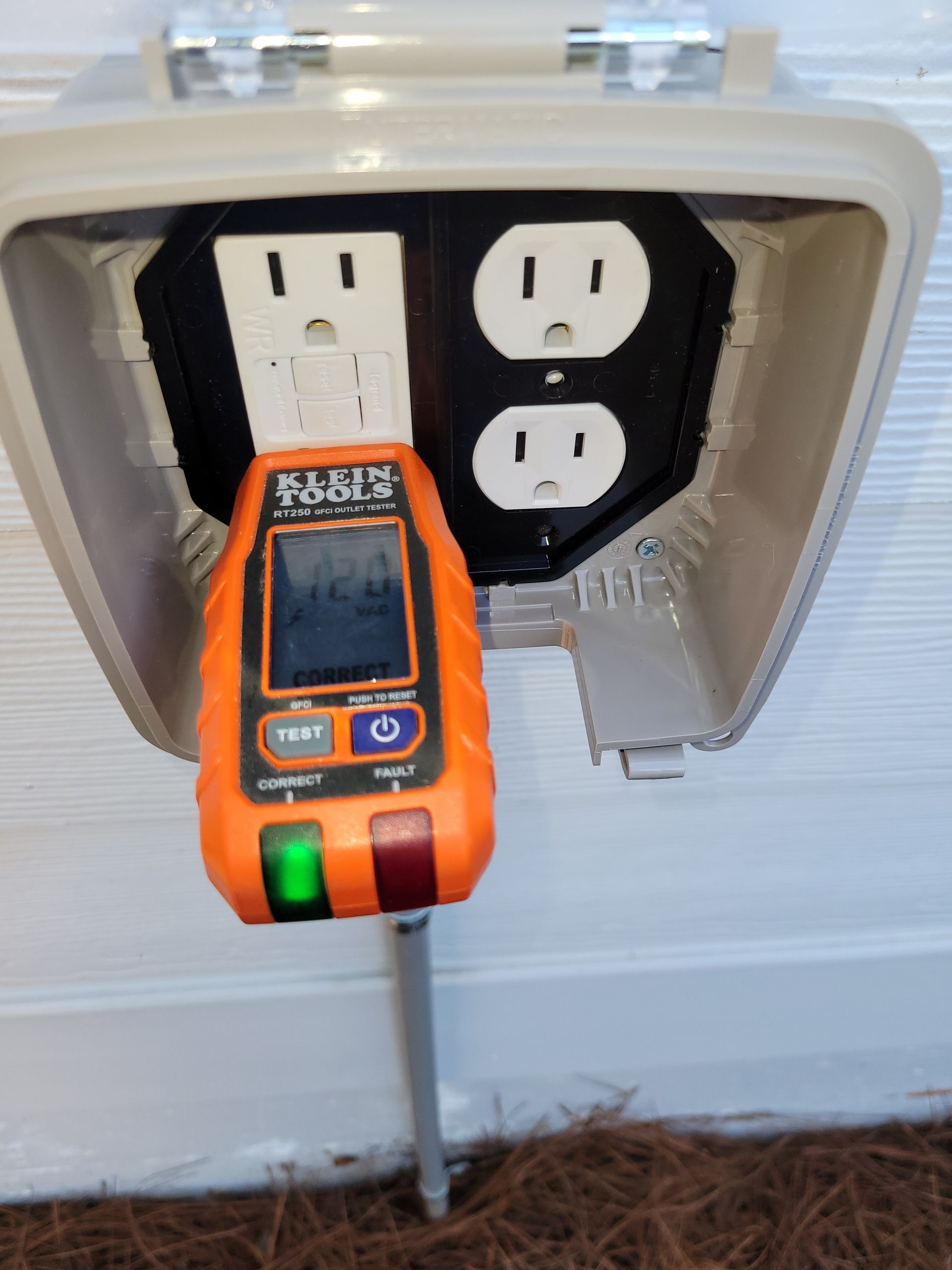 Orange electrical tester in outdoor outlet box, testing outlet. Green and red indicator lights are lit.