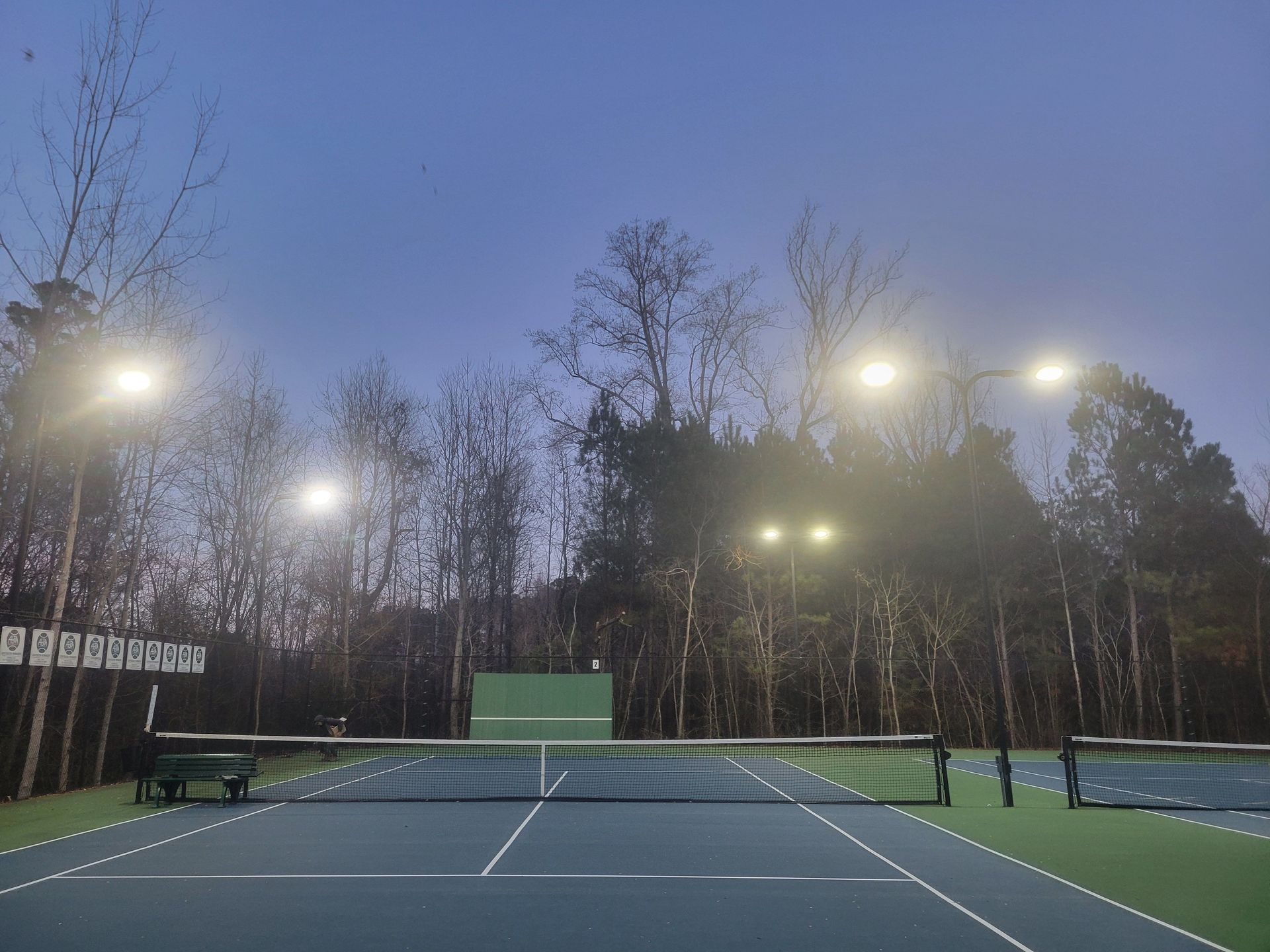 Tennis court at dusk with illuminated lights and trees in the background.
