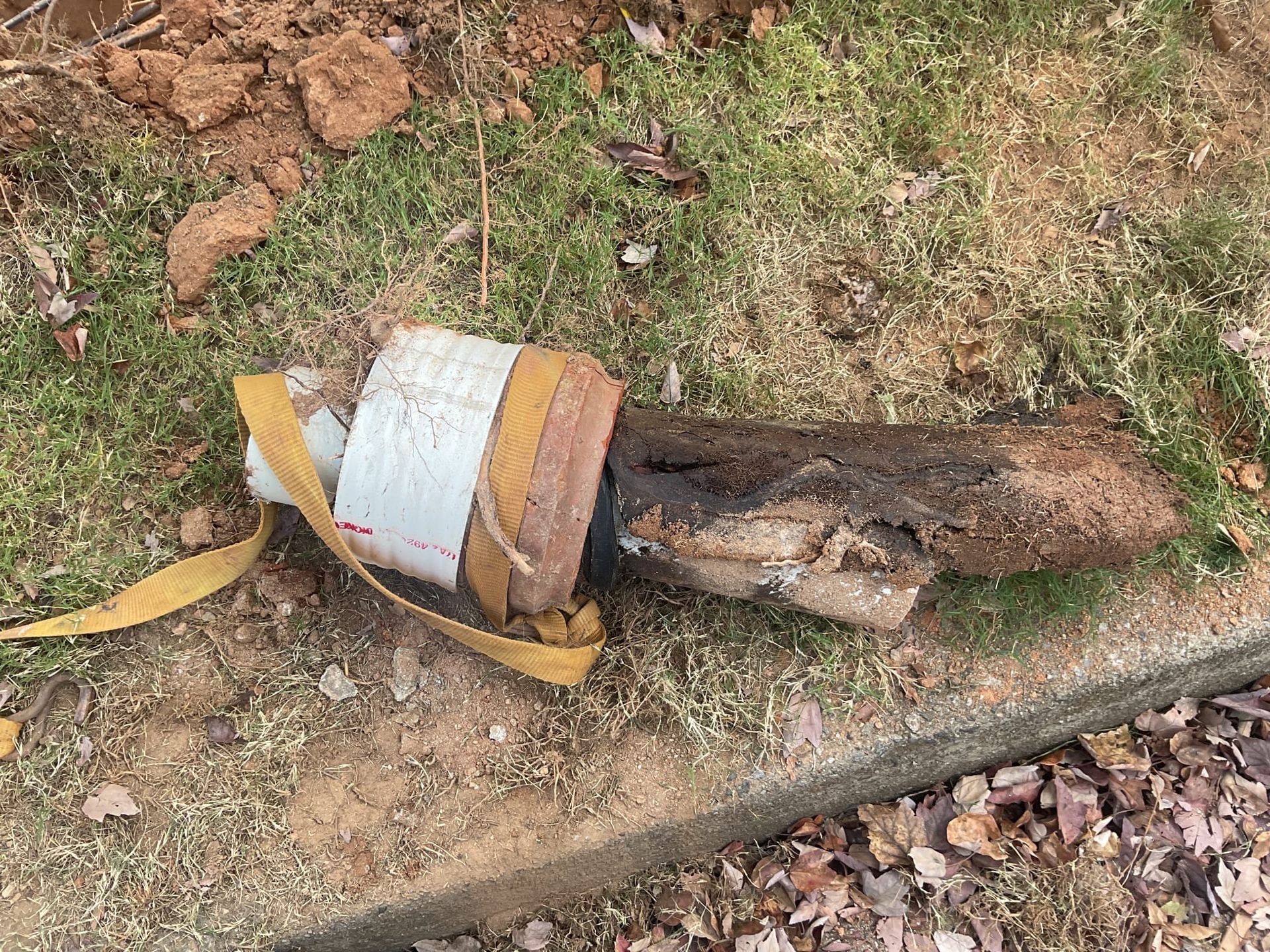 White wrapped pipe with yellow strap next to a brown, decaying log on grass and dirt.