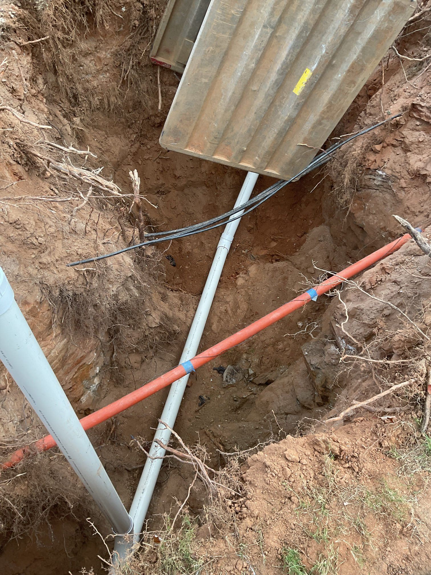 Trench with exposed pipes and wires, partially covered by corrugated metal. Earth tones dominate.