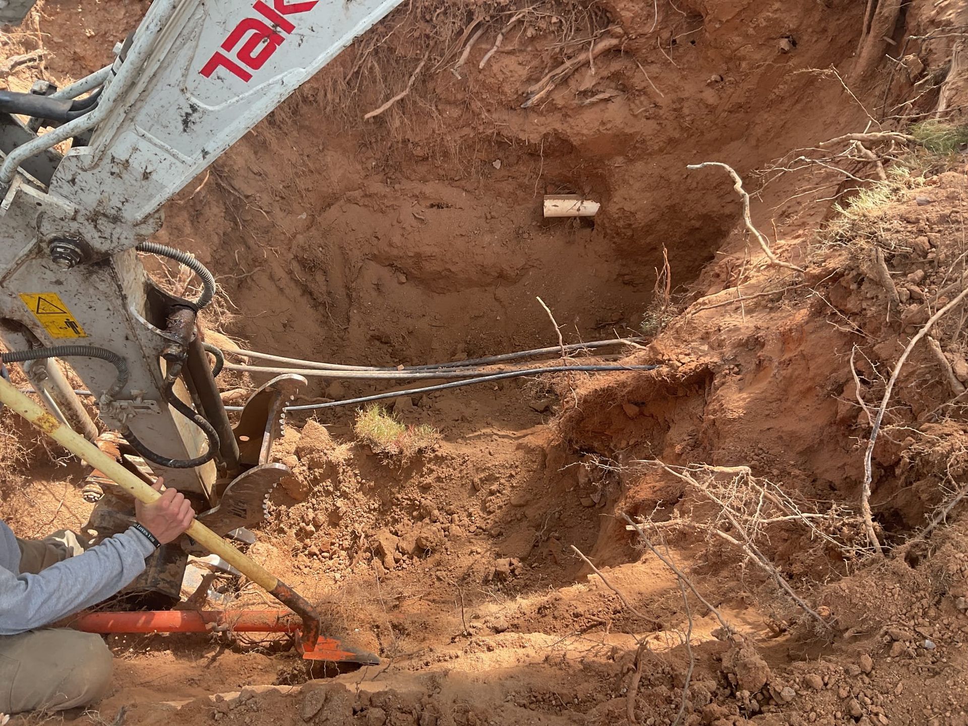 Excavator digging into the earth near a person using a shovel. Visible are pipes and exposed wires.