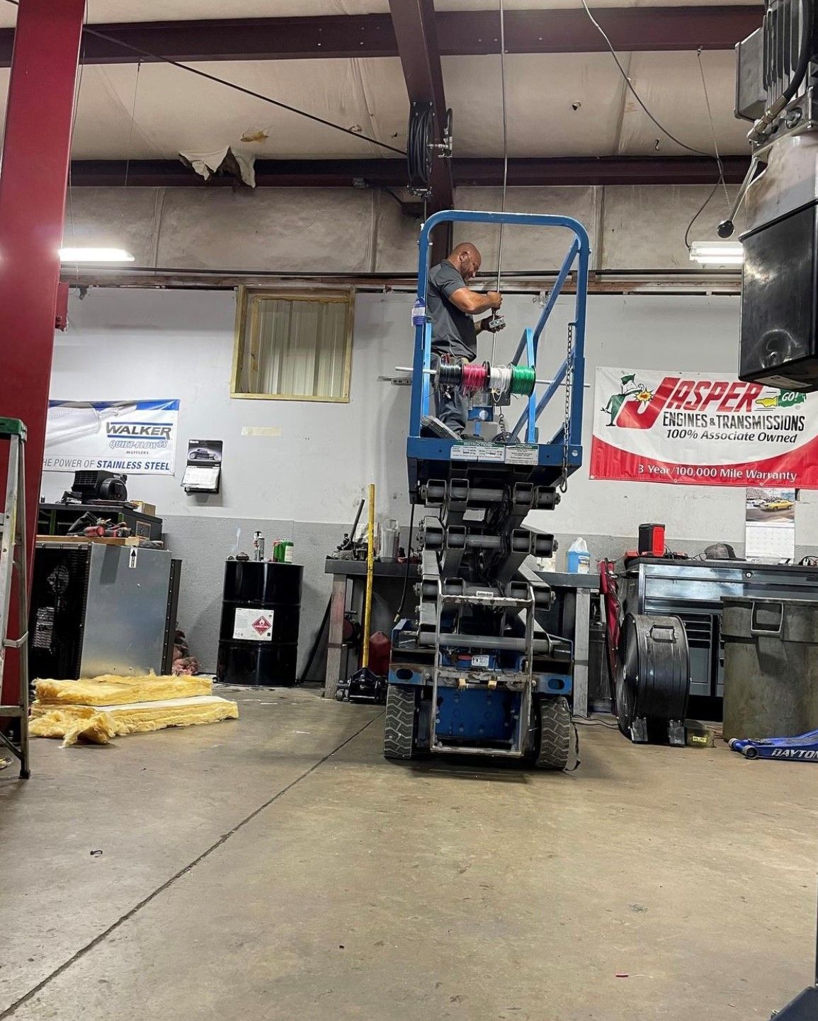 Man operating a blue lift inside a garage. He’s at a height, seemingly working on something overhead.