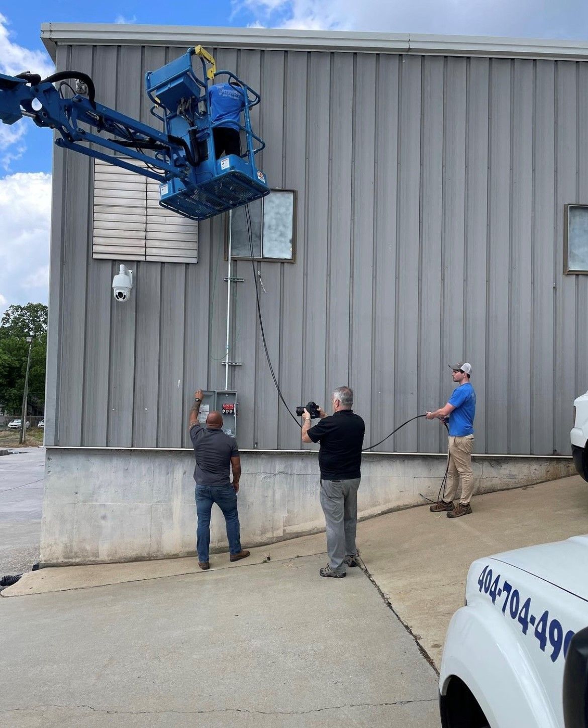 Workers using a lift to access a building's electrical panel. Two workers on the ground, one using a cable puller.