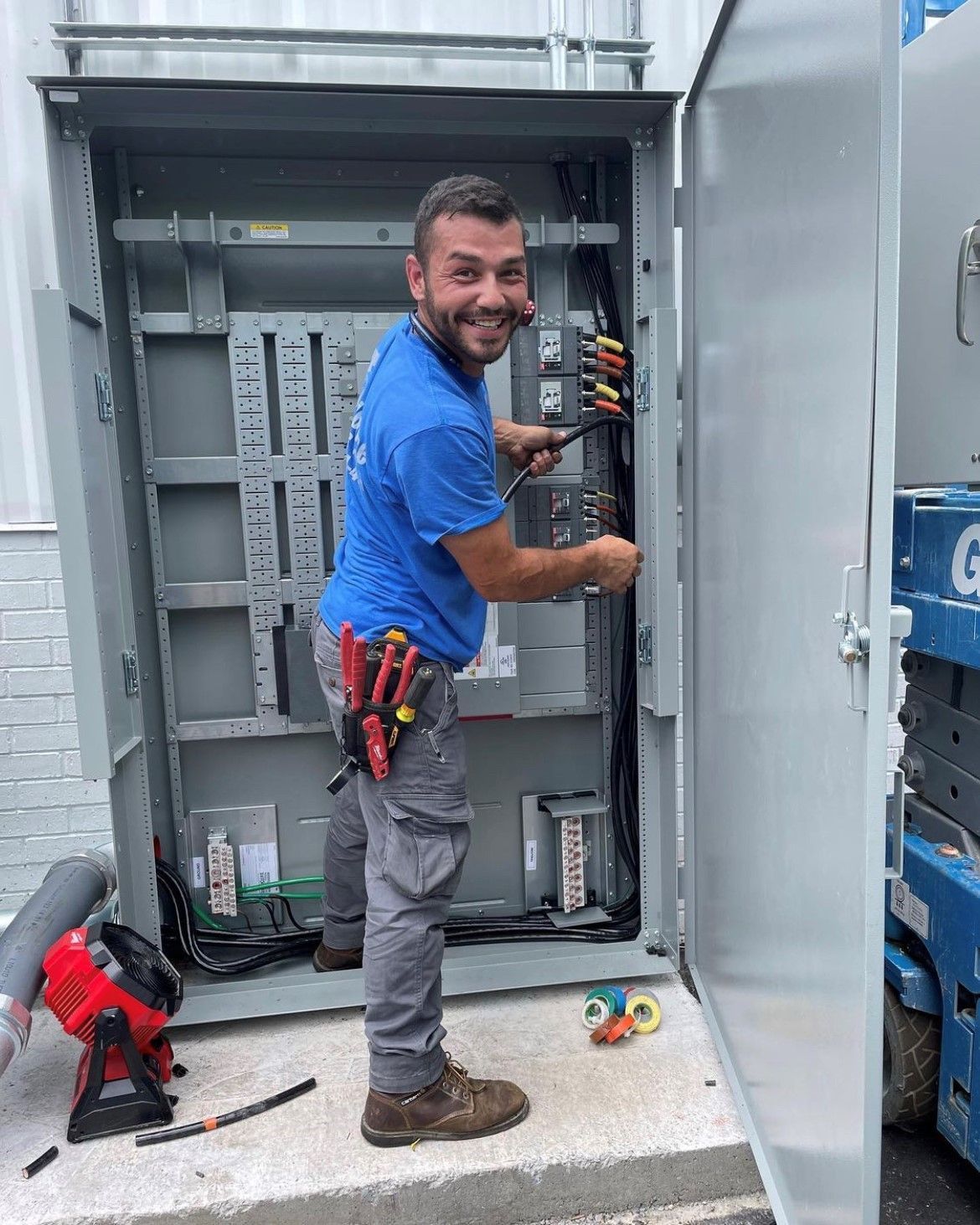 Electrician working inside an electrical cabinet, smiling. Wearing blue shirt, gray pants, and tools. Outdoors, near equipment.