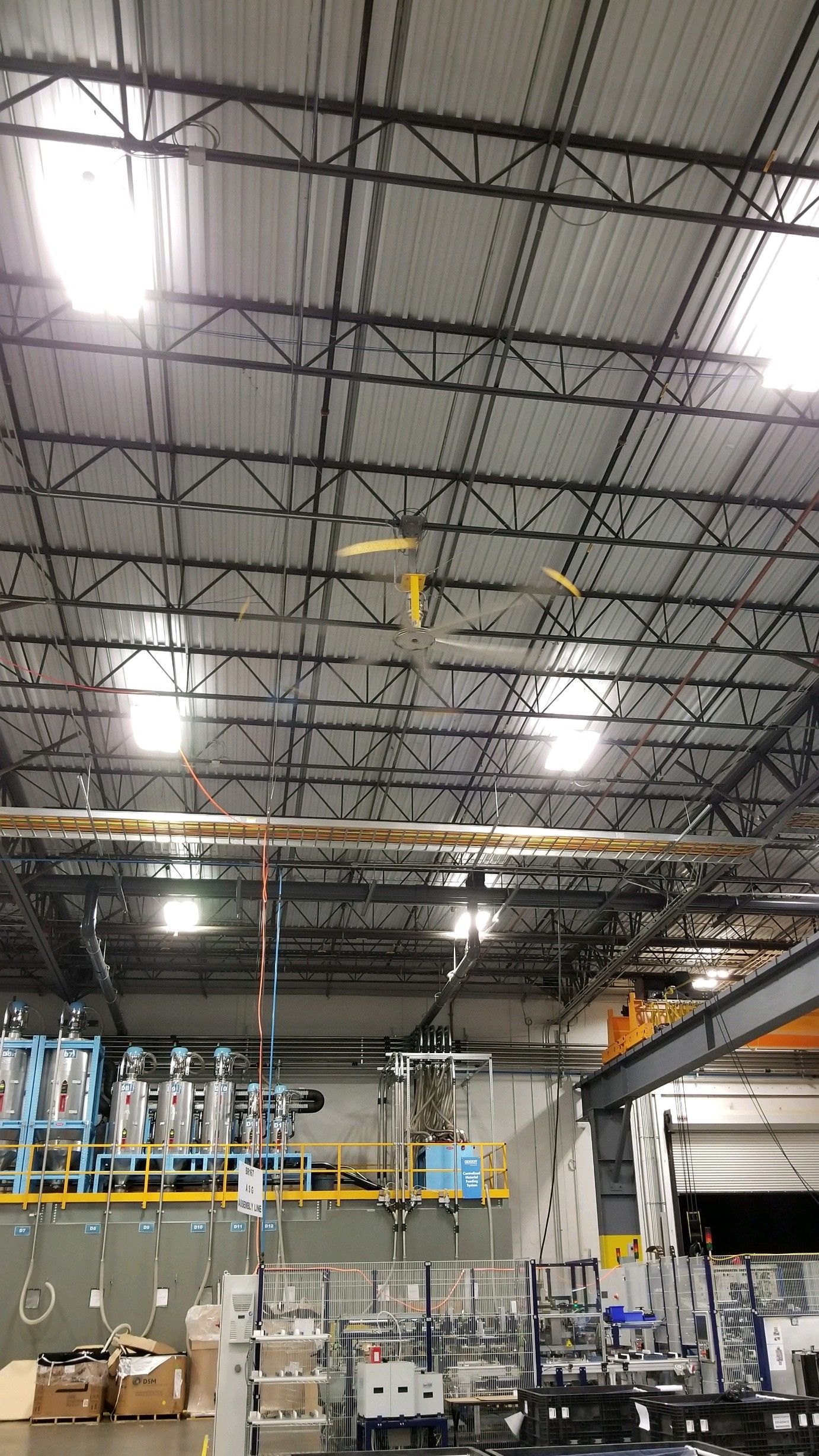 Industrial factory interior with a high ceiling, overhead lighting, and machinery. Yellow fan blades are visible.