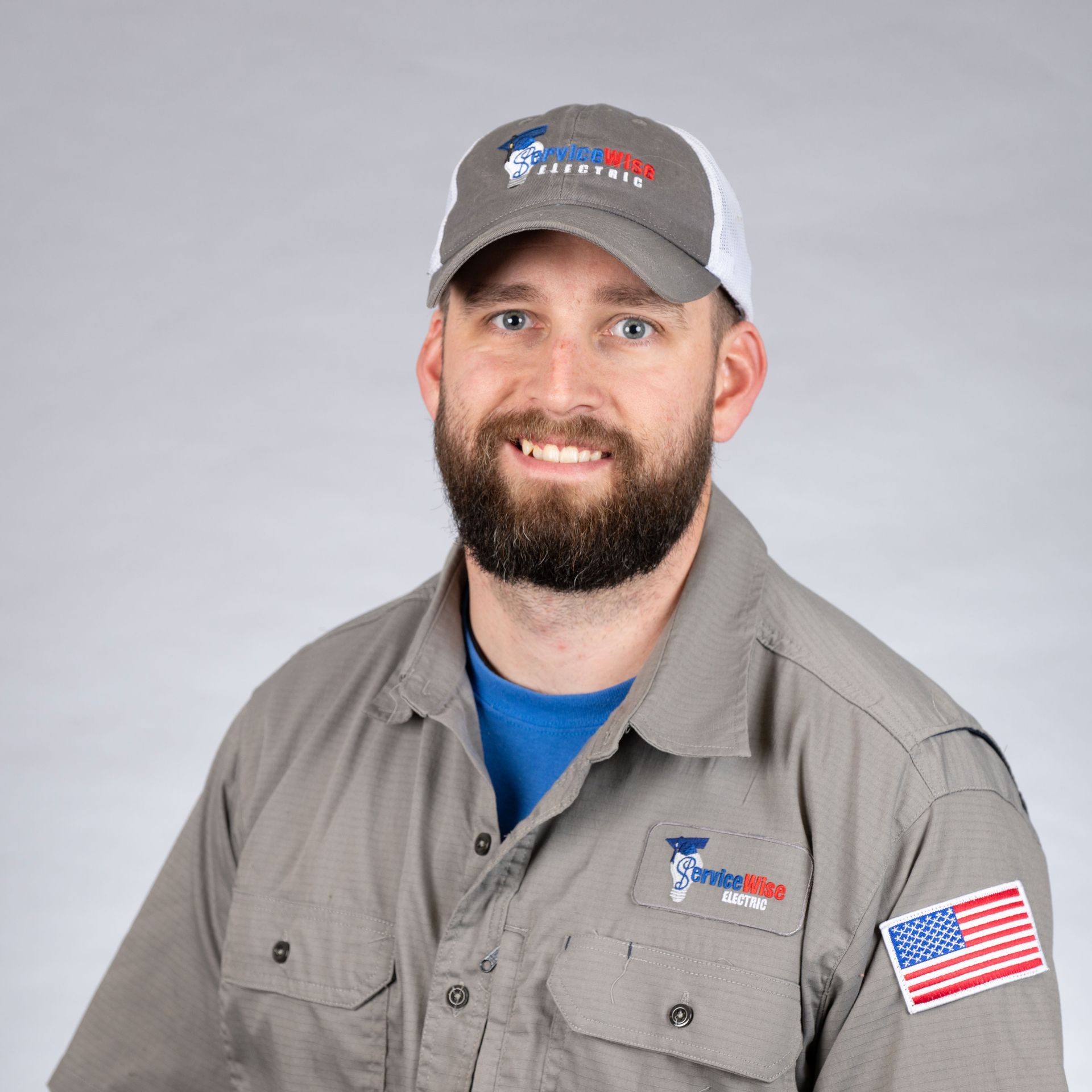 Man in grey work shirt and cap, smiling, with the US flag patch on the sleeve.