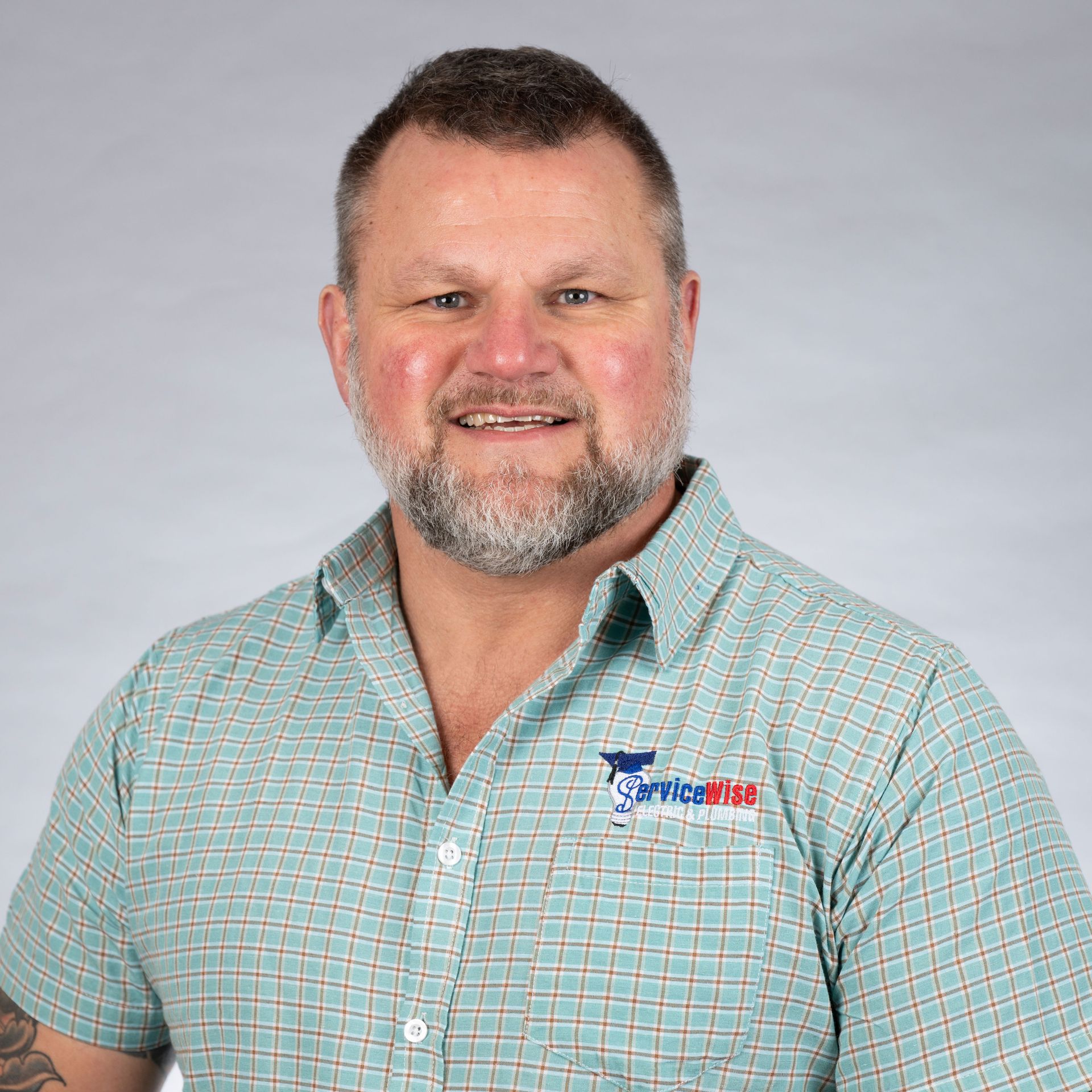 Man with a beard, wearing a green plaid shirt with a logo, smiling, headshot.