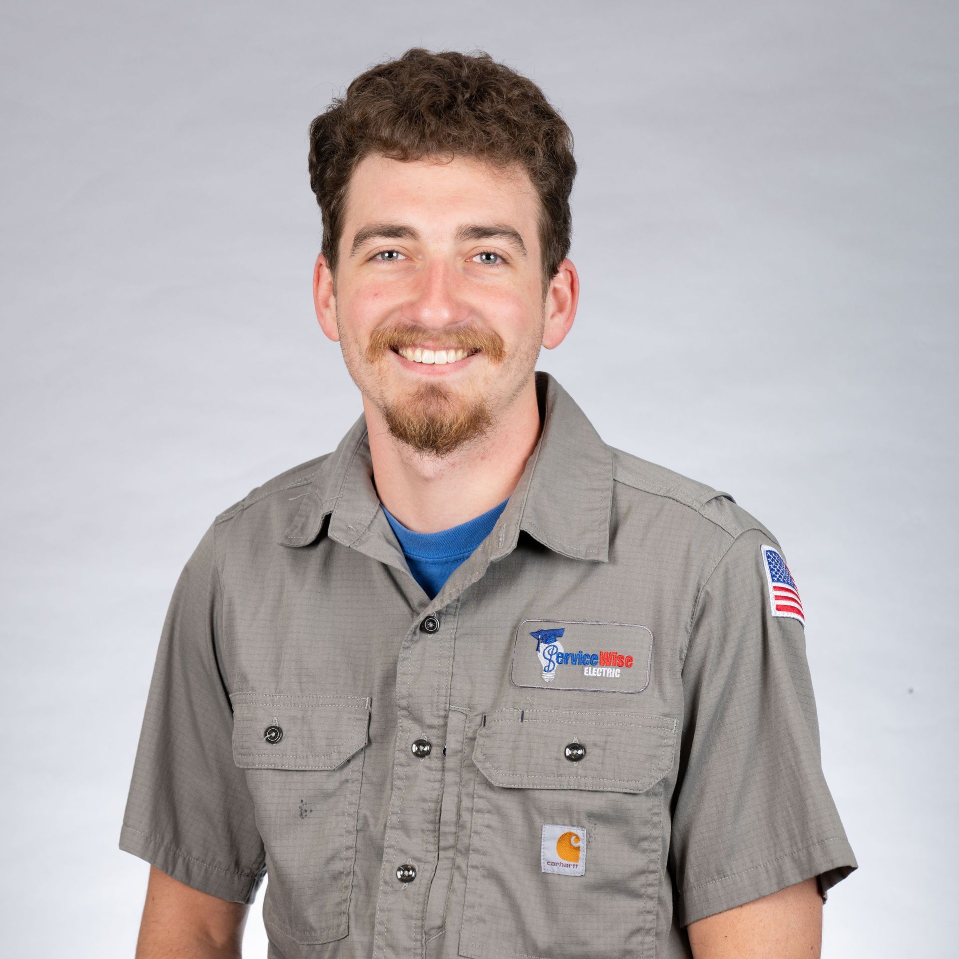 Man in gray work shirt smiles, with beard, outdoors. American flag patch on sleeve.