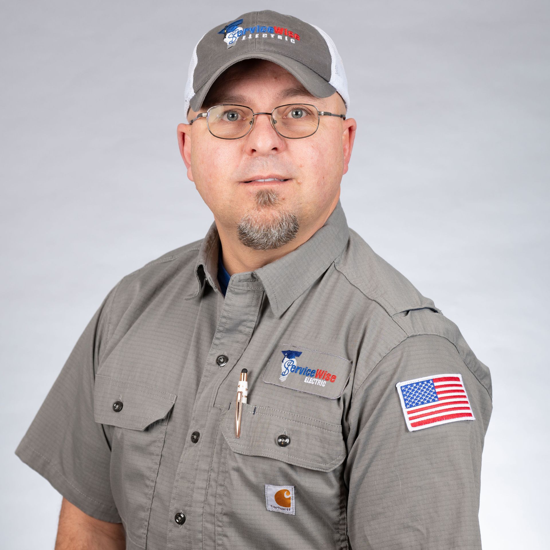 Man in grey work shirt and cap, holding pen, with American flag patch.