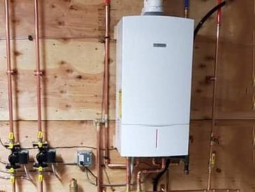 A white water heater is hanging on a wooden wall next to copper pipes.