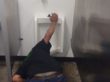 A man is laying on the floor in a public restroom next to a urinal.