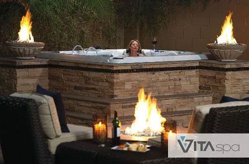 Woman relaxes in a hot tub with fire features in a backyard setting.