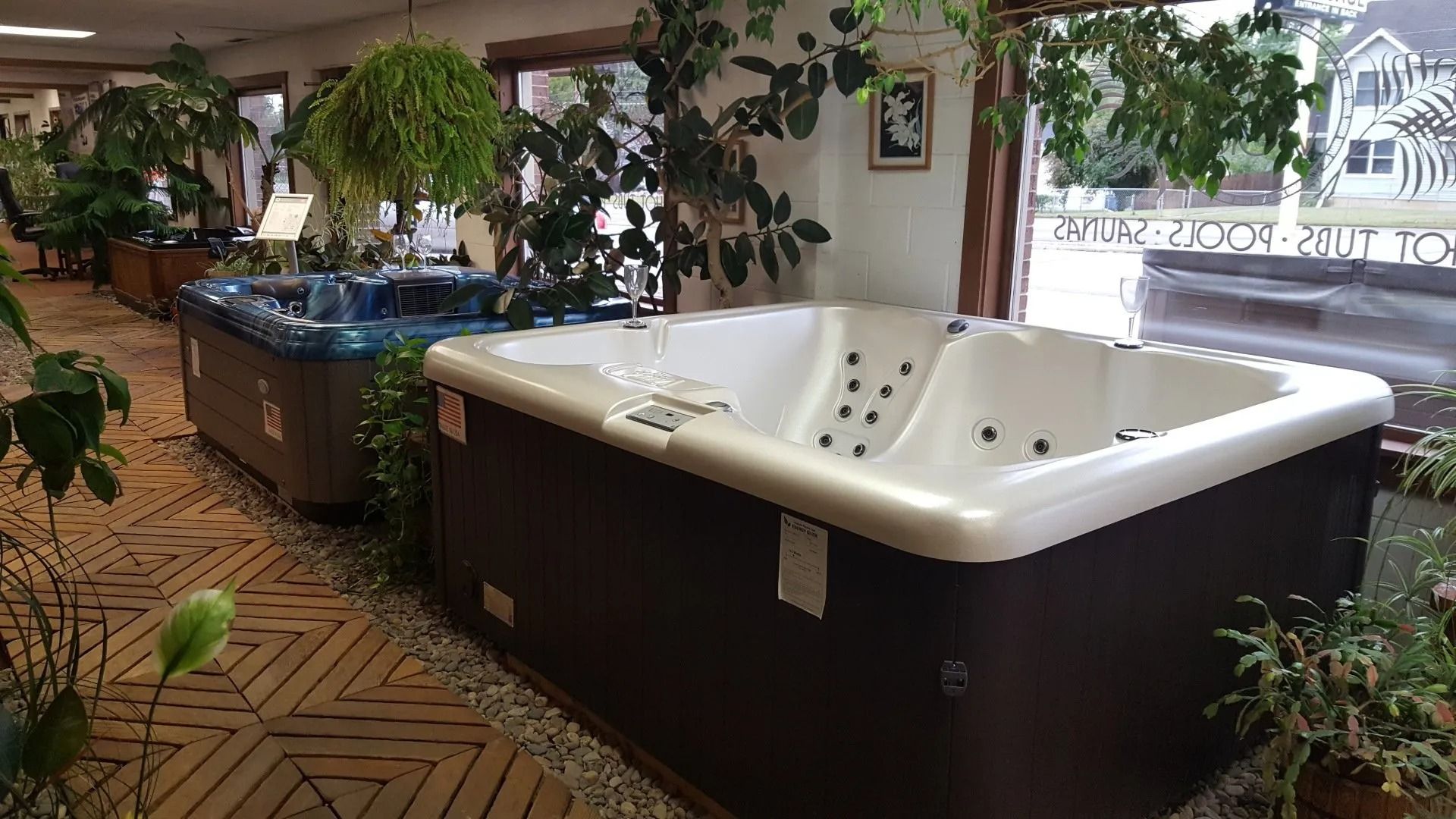 Indoor showroom with two hot tubs, plants, and a window. One hot tub is white, the other blue.