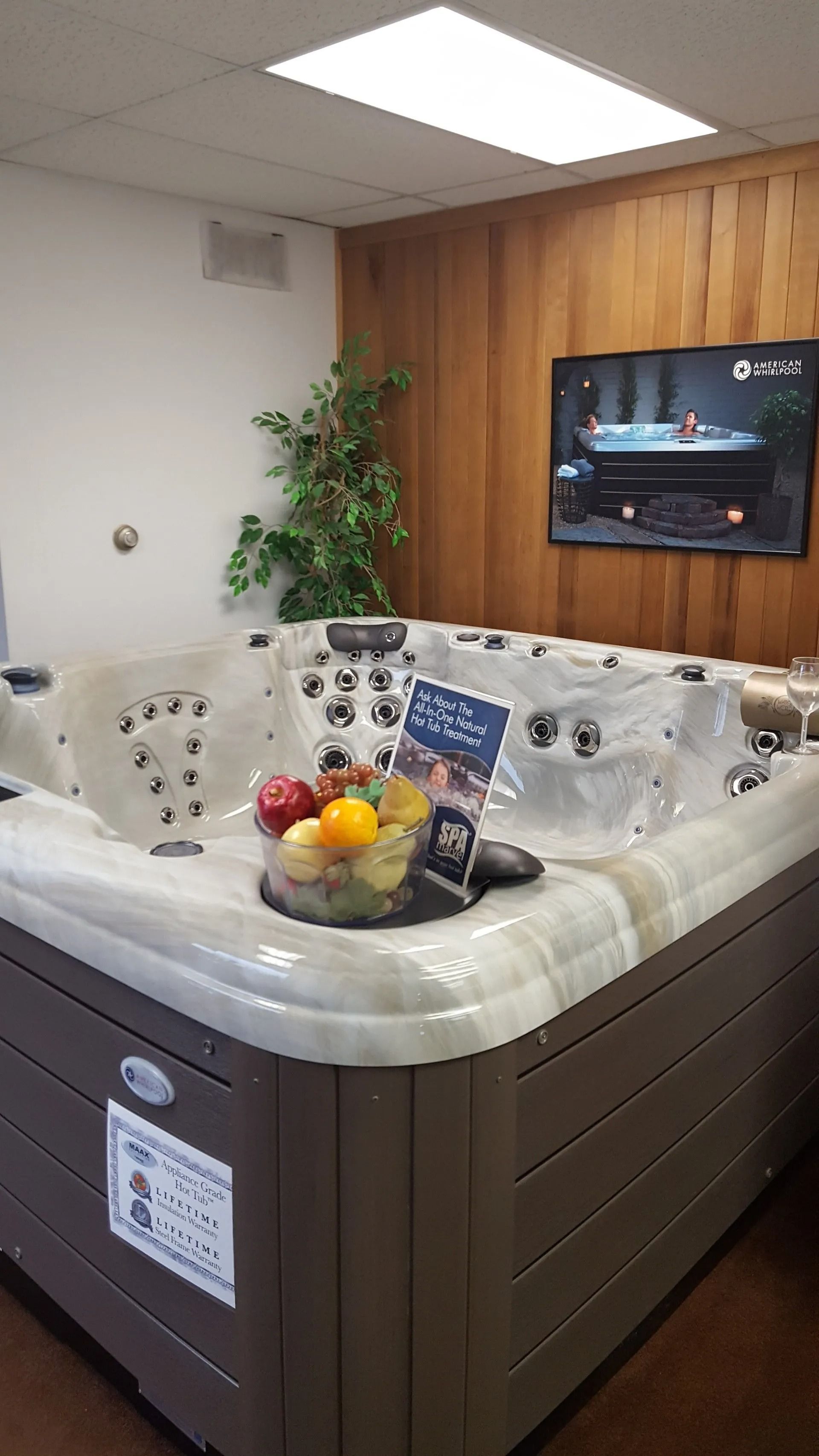 Hot tub on display in a store; fruit bowl on the edge, wooden paneling, TV, and a green plant.