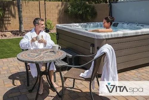 A man and woman relax near a hot tub outdoors. The man in a robe sips from a mug. The woman sits in the tub.