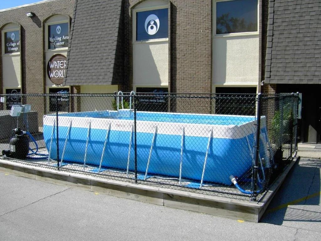 Blue above-ground pool enclosed by a black fence outside a brick building with logo.