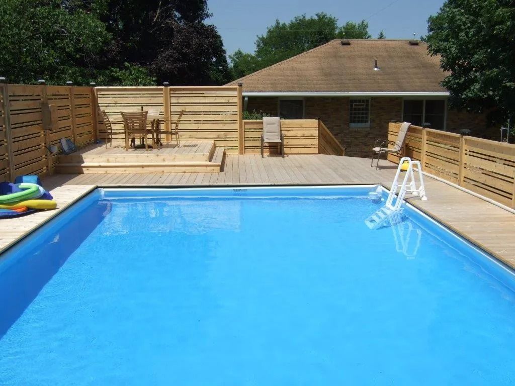 A rectangular pool with wooden deck, chairs, and horizontal fence in backyard. House in background.