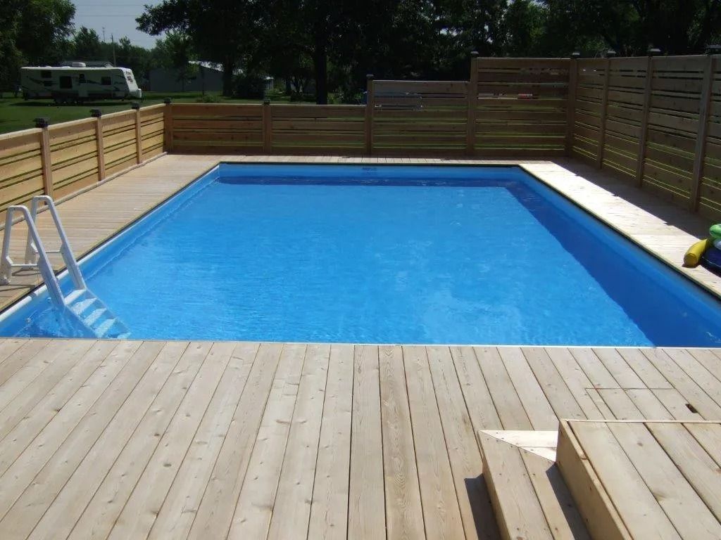 Rectangular swimming pool surrounded by a wooden deck and fence. The pool water is blue.
