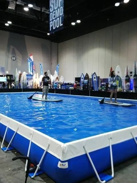 People paddleboarding in a blue indoor demo pool at a trade show, surrounded by surfboards and equipment.