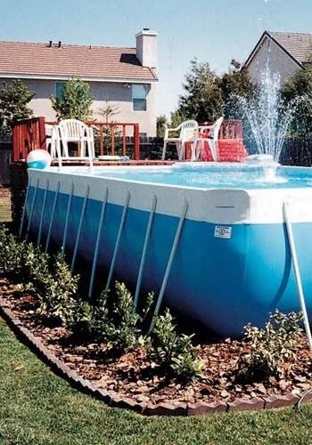 Above-ground blue pool with fountain, white chairs on deck, surrounded by shrubs and lawn.