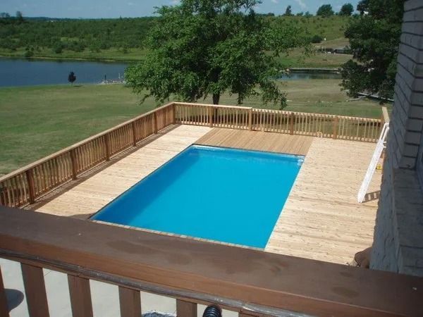 Above-ground pool surrounded by wooden deck and railing, overlooking a lake and green landscape.