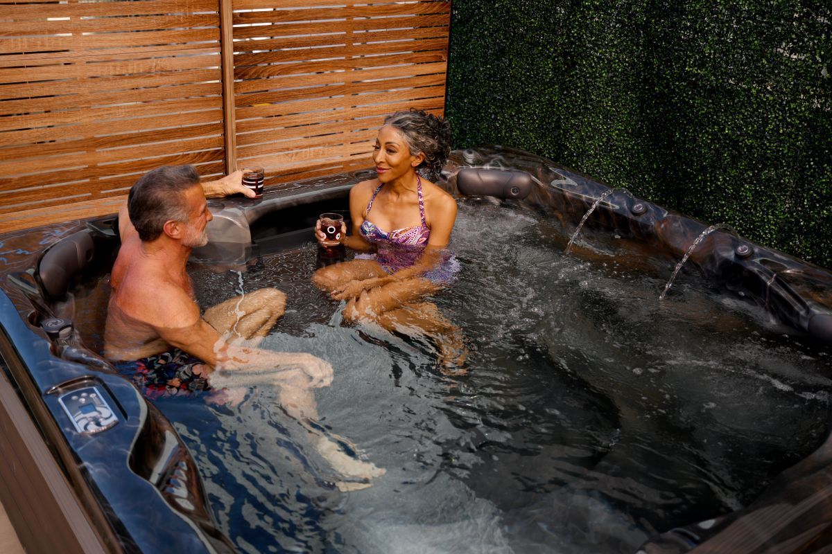 Couple relaxing in a hot tub, holding drinks, with wooden fence and green wall backdrop.