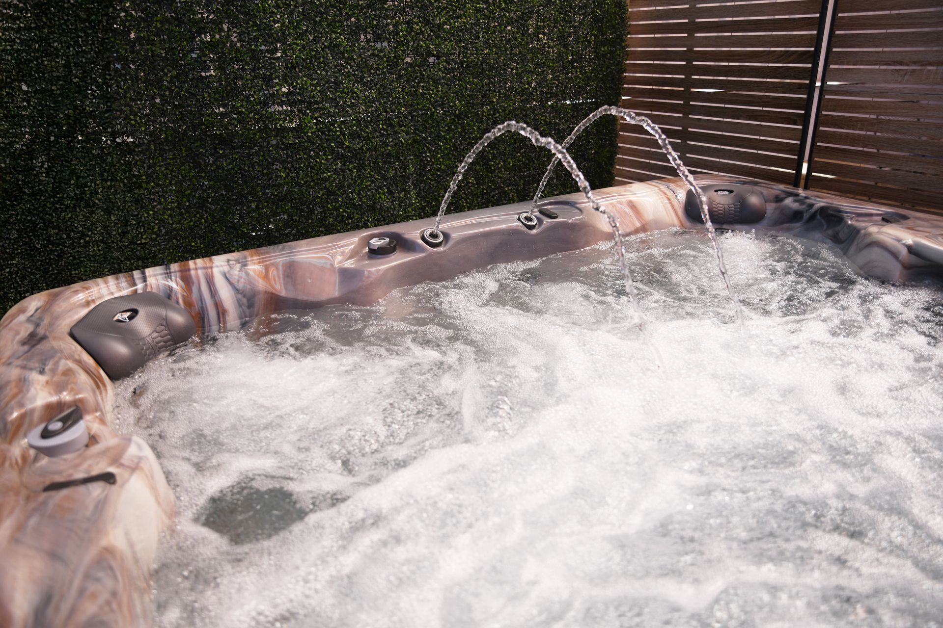 Hot tub with jets spraying water; green hedge and brown fence in background.