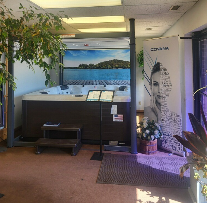 Hot tub display in a showroom, with a lake scene backdrop and "COVANA" banner. Brown tub with steps.