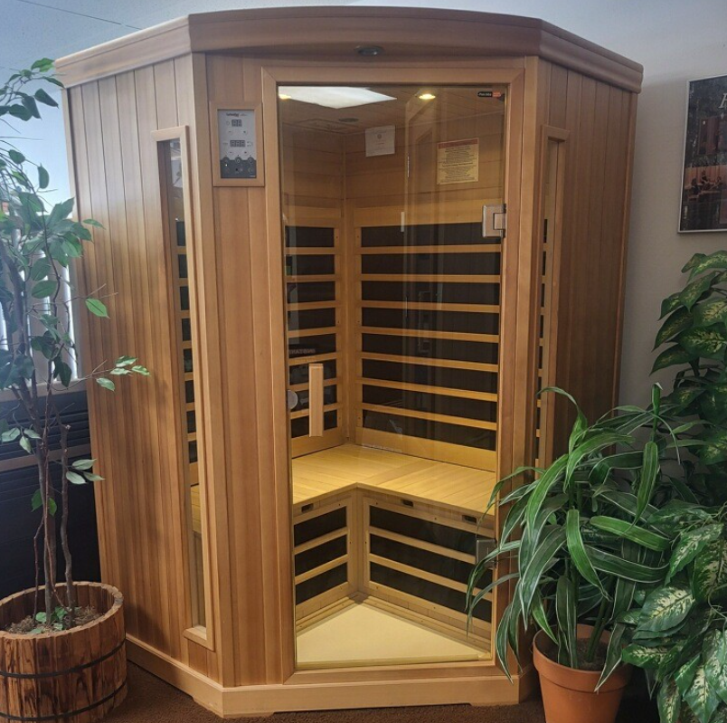 Wooden corner infrared sauna with glass door and side window; plants in the foreground.