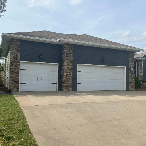 Two-car garage with white doors, gray siding, stone accents, and concrete driveway against a cloudy sky.