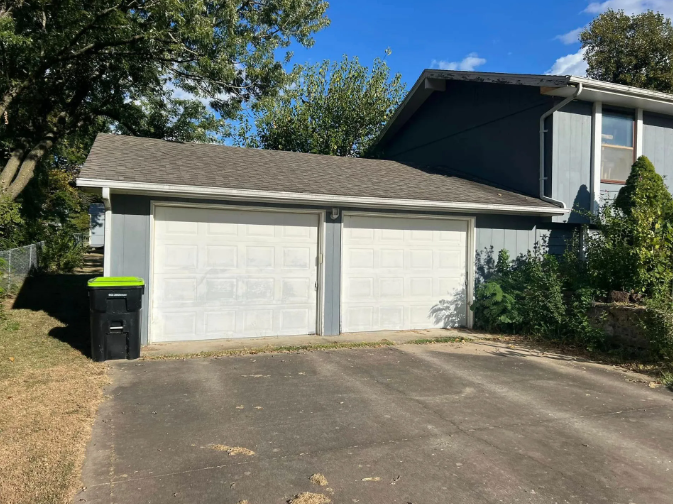 Two-car garage with white doors, gray siding, and a black trash can on a driveway.