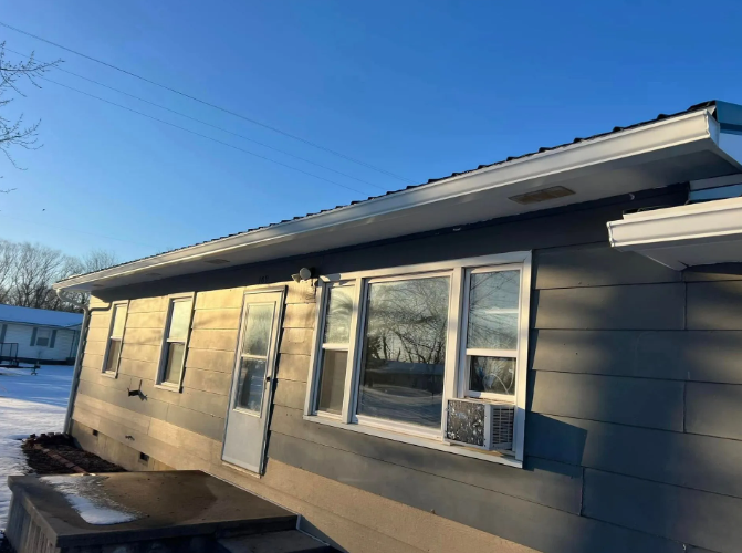 A single-story house with gray siding and white trim. Windows and a door are visible. Snow and blue sky.