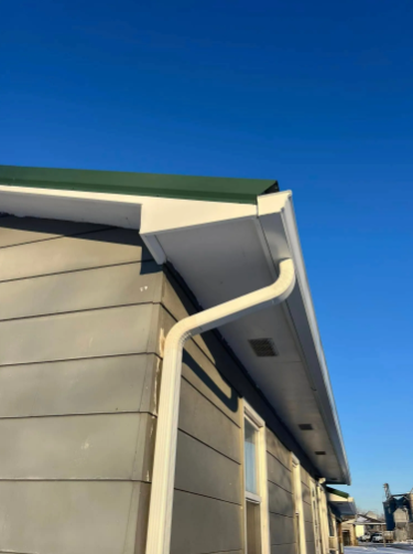 White gutter along the edge of a gray building with a green roof, against a clear blue sky.
