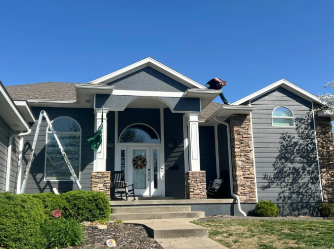 Person on a roof working on gutters of a blue house with stone accents and a white front door.