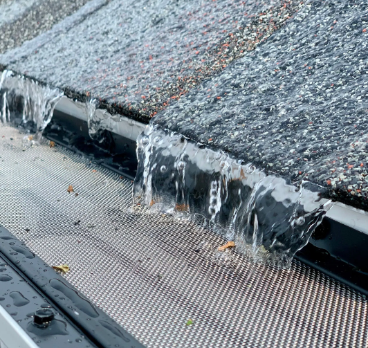 Water flowing over a partially blocked gutter with a mesh screen, roof in the background.