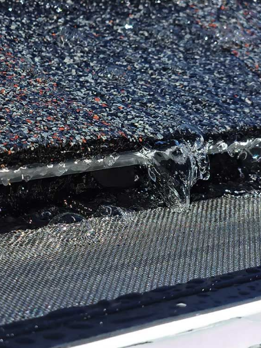 Water cascading from a clogged gutter onto a mesh screen; shingles in background.