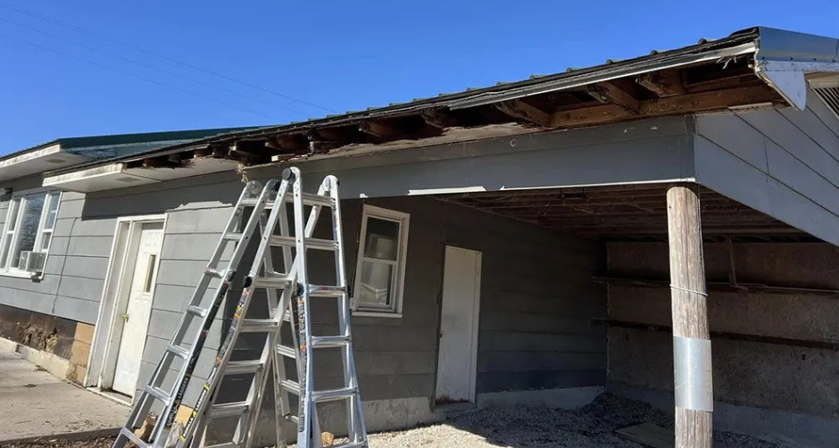 Damaged house exterior with exposed roof under repair; ladder propped up.
