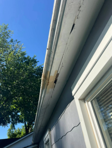 Damaged white soffit with cracks and discoloration, likely from water damage, near a window. Blue sky and tree visible.