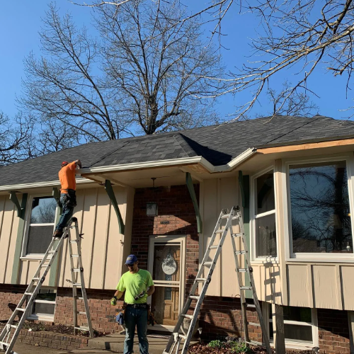 Two workers installing a roof on a house with ladders, clear sky.