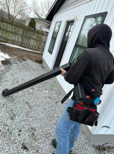 Person carrying a black gutter piece next to a white house with black window frames, gravel ground.