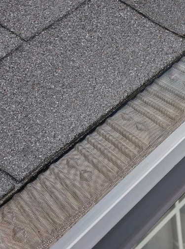 Water overflowing from a gutter onto the side of a roof with dark gray shingles.