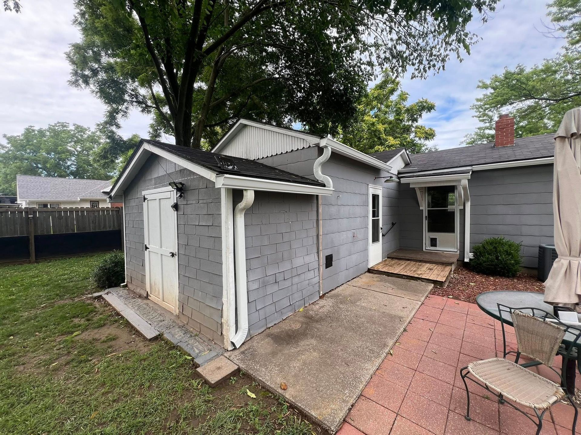 Gray shed and house connected by a concrete walkway. Red brick patio has outdoor furniture.