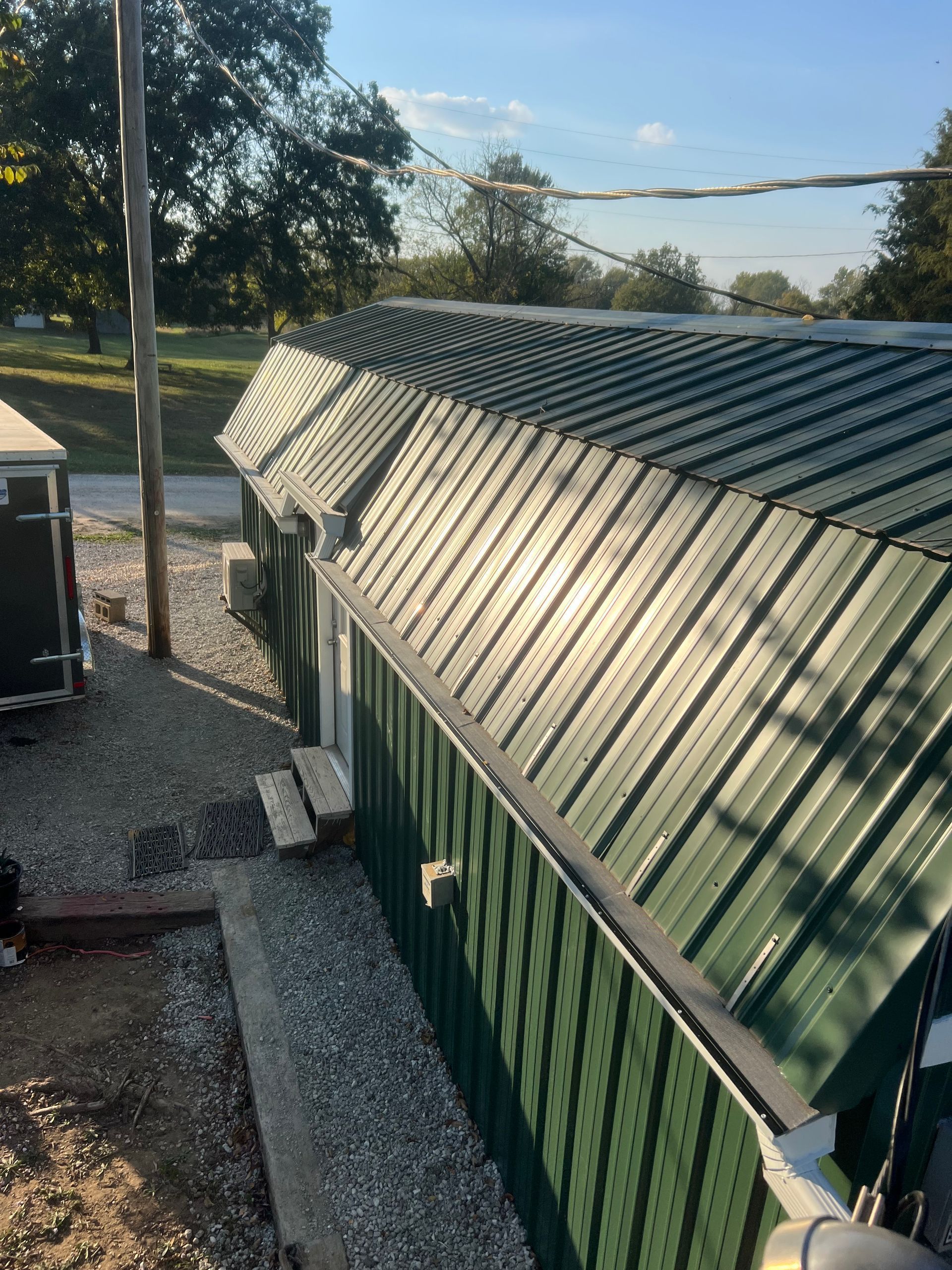An elevated angle shows a dark green metal building with a gambrel-style roof, set on a gravel lot near trees.
