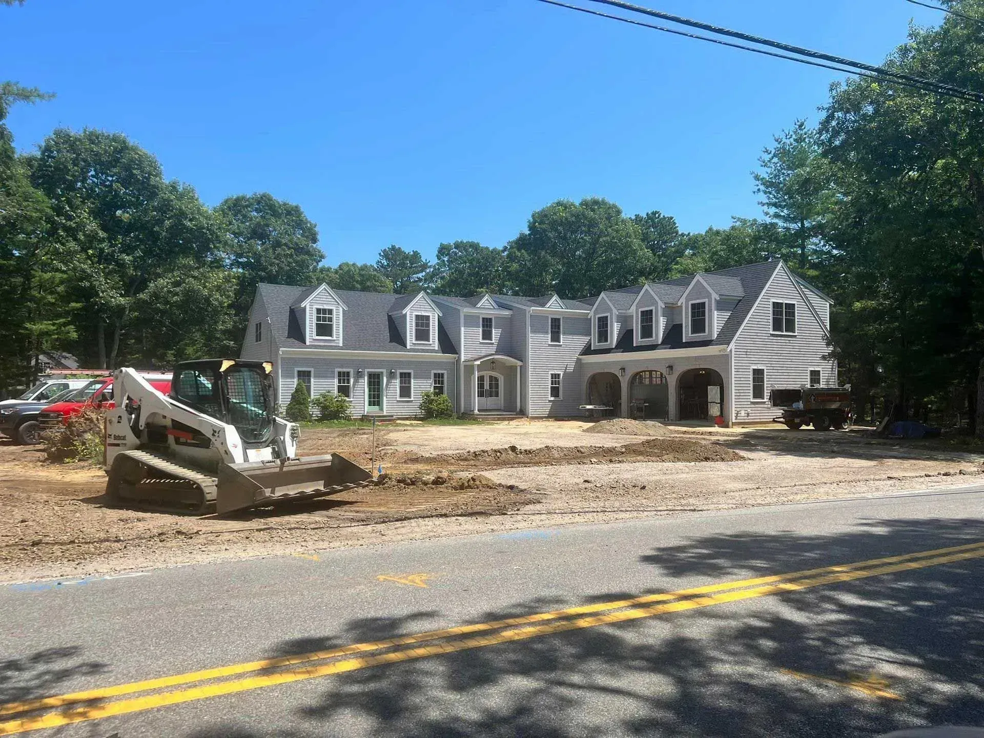 Construction equipment clearing dirt in front of a white house on a tree-lined roadside