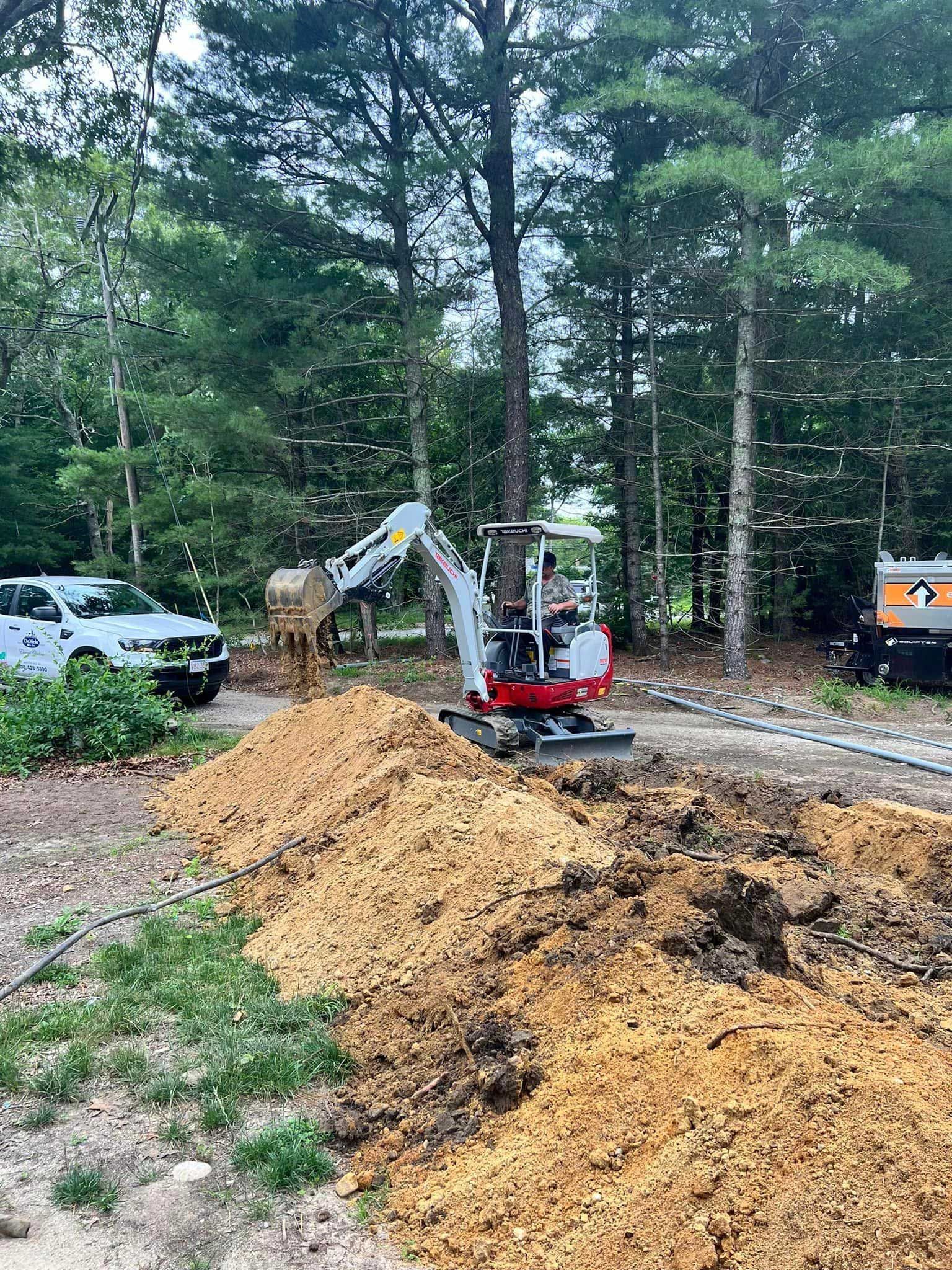 Mini excavator digging soil beside a wooded road, with a white truck and orange construction equipment nearby